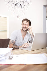 Smiling man sitting at wooden table with laptop and folder telephoning with smartphone - MFRF000426