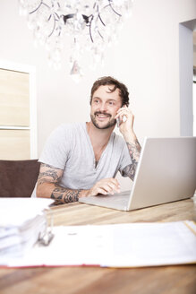Lächelnder Mann sitzt an einem Holztisch mit Laptop und Ordner und telefoniert mit einem Smartphone - MFRF000426