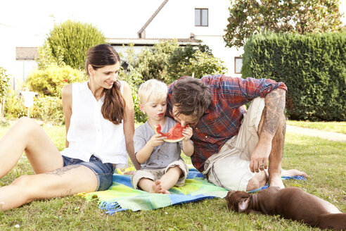 Eltern sitzen mit ihrem kleinen Sohn auf einer Decke im Garten und essen Wassermelone - MFRF000433