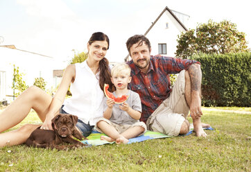 Parents sitting with their little son and dog on a blanket in the garden - MFRF000434