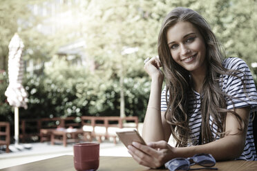 Portrait of smiling young woman sitting at a table holding smartphone - GDF000872