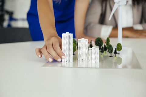 Nahaufnahme einer Hand, die ein Hochhausmodell neben einem Windradmodell auf einem Konferenztisch hält, lizenzfreies Stockfoto