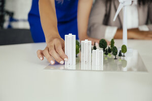 Close-up of hand holding high-rise building model next to wind turbine model on conference table - MFF002148