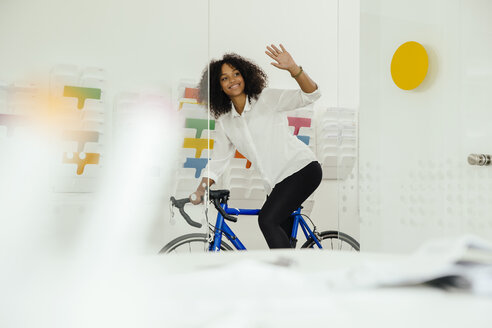 Lächelnde junge Frau mit Fahrrad im Büro winkt - MFF002179