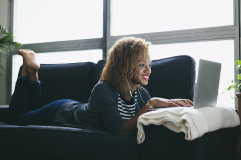 Junge Frau auf der Couch liegend mit Kopfhörern und Laptop, lizenzfreies Stockfoto