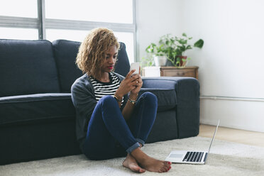 Junge Frau sitzt auf dem Teppich im Wohnzimmer und schaut auf ihr Smartphone - EBSF000887