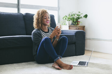 Lächelnde junge Frau sitzt auf dem Teppich im Wohnzimmer und schaut auf ihr Smartphone - EBSF000888