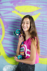Porträt eines lächelnden Teenager-Mädchens, das ein buntes Skateboard vor einer Wand mit Graffiti hält - GEMF000371