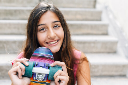 Porträt eines lächelnden Teenager-Mädchens mit buntem Skateboard auf einer Treppe sitzend - GEMF000376