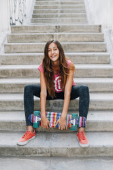 Portrait of happy teenage girl with colorful skateboard sitting on stairs - GEMF000377