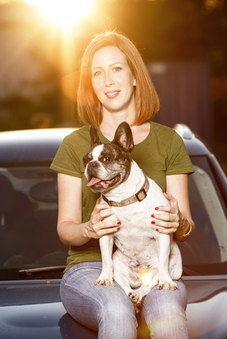 Portrait of redheaded woman sitting on car bonnet with her French bulldog stock photo