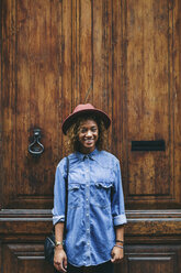 Spanien, Barcelona, Porträt einer lächelnden jungen Frau mit Hut und Jeanshemd, die vor einer Holztür steht - EBSF000912