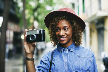 Porträt einer lächelnden jungen Frau mit Hut und Jeanshemd, die eine Kamera hält - EBSF000919