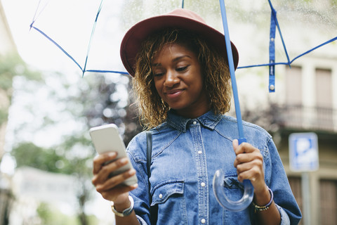 Spanien, Barcelona, Porträt einer lächelnden jungen Frau mit Regenschirm und Smartphone, lizenzfreies Stockfoto