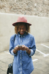 Porträt einer lächelnden jungen Frau mit Hut und Jeanshemd, die auf ihr Smartphone schaut - EBSF000928