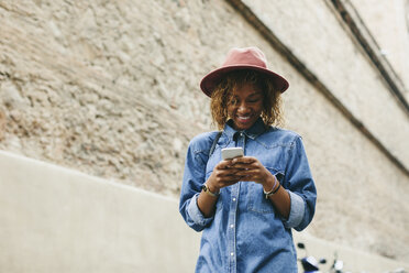 Porträt einer lächelnden jungen Frau mit Hut und Jeanshemd, die auf ihr Smartphone schaut - EBSF000929