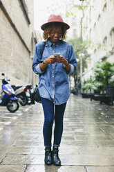 Spain, Barcelona, portrait of young woman wearing hat and denim shirt looking at her smartphone - EBSF000930