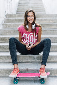 Portrait of teenage girl sitting on stairs with a skateboard and headphones - GEMF000383