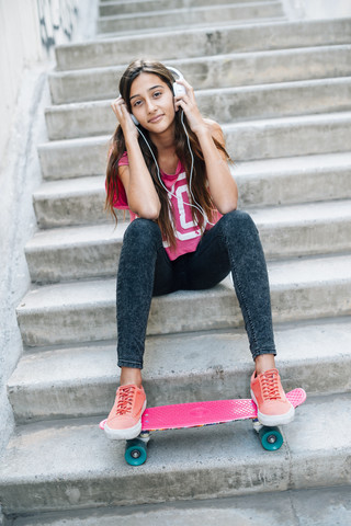 Porträt eines Teenagers, der auf einer Treppe mit einem Skateboard sitzt und mit Kopfhörern Musik hört, lizenzfreies Stockfoto