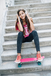 Porträt eines Teenagers, der auf einer Treppe mit einem Skateboard sitzt und mit Kopfhörern Musik hört - GEMF000382