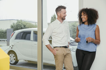 Mann und Frau unterhalten sich beim Aufladen eines Elektroautos, Frau hält Schlüssel - MFF002216