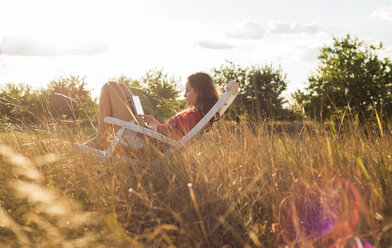 Woman sitting on a deck chair on a meadow using digital tablet - UUF005646