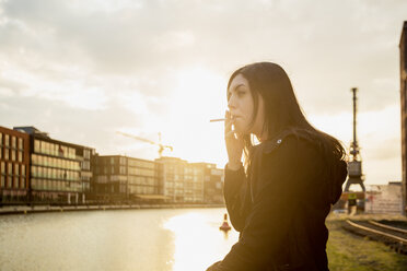 Germany, Muenster, young woman smoking cigarette in front of city harbour - TAM000299