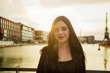 Germany, Muenster, portrait of young woman standing in front of city harbour at backlight - TAM000297