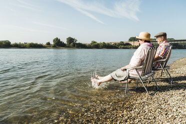 Germany, Ludwigshafen, senior couple sitting on folding chairs at riverside splashing with feet in water - UUF005680