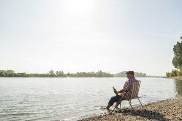 Deutschland, Ludwigshafen, älterer Mann mit Bierflasche auf Klappstuhl am Flussufer sitzend - UUF005713