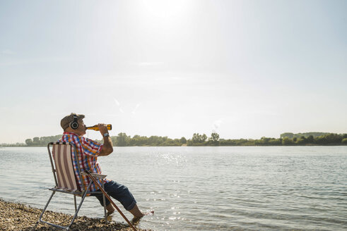 Deutschland, Ludwigshafen, älterer Mann mit Kopfhörern sitzt auf einem Klappstuhl am Flussufer und trinkt Bier - UUF005682