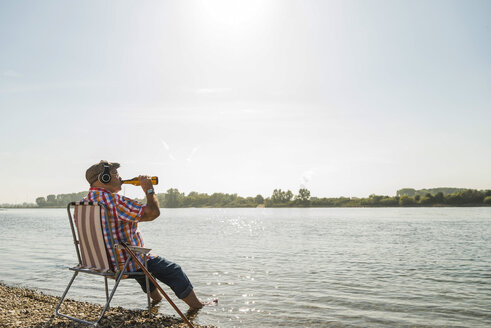 Germany, Ludwigshafen, senior man with headphones sitting on folding chair at riverside drinking beer - UUF005682