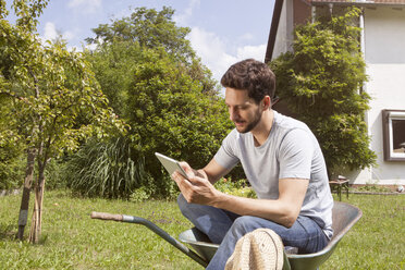 Man sitting in wheelbarrow in garden using digital tablet - RBF003147