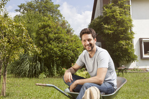 Lächelnder Mann sitzt in einer Schubkarre im Garten, lizenzfreies Stockfoto