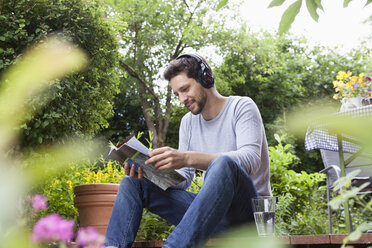 Relaxed man sitting in garden with headphones and magazine - RBF003205