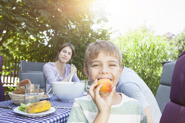 Smiling boy with his family holding a fruit at garden table - RBF003186
