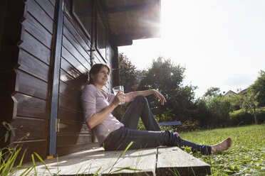 Smiling woman sitting at garden shed with glass of water - RBF003197