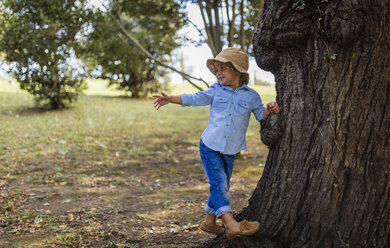Porträt eines lächelnden blonden kleinen Jungen mit Hut, der neben einem Baum steht - MGOF000769