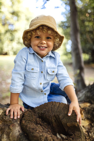 Porträt eines lächelnden blonden kleinen Jungen mit Hut, der auf totem Holz sitzt, lizenzfreies Stockfoto