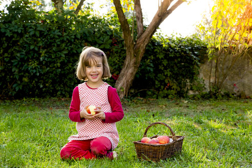 Portrait of smiling little girl sitting on a meadow with wickerbasket of apples - LVF003868