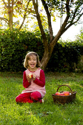 Portrait of laughing little girl sitting on a meadow with wickerbasket of apples - LVF003869