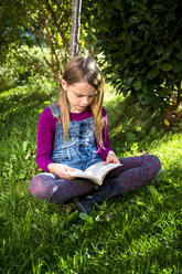 Little girl sitting on a meadow in the garden reading a book - SARF002111