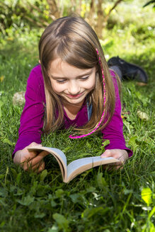 Happy little girl lying on a meadow in the garden reading a book - SARF002112
