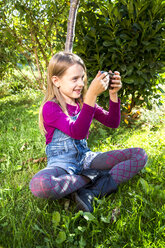 Little girl sitting on a meadow in the garden taking a selfie with smartphone - SARF002114