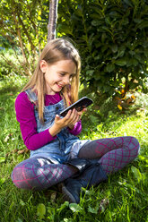 Kleines Mädchen sitzt auf einer Wiese im Garten und schaut auf ihr Smartphone - SARF002115