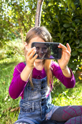 Little girl sitting on a meadow in the garden showing display of smartphone with her selfie - SARF002117