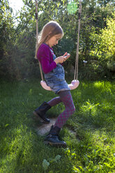 Little girl sitting on a swing in the garden looking at smartphone - SARF002119