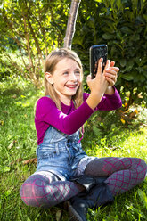 Kleines Mädchen sitzt auf einer Wiese im Garten und macht ein Selfie mit Smartphone - SARF002120