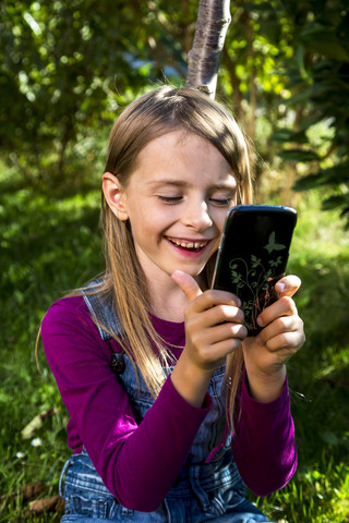 Glückliches kleines Mädchen betrachtet Smartphone in einem Garten, lizenzfreies Stockfoto