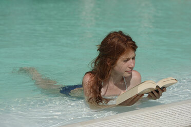 Redheaded girl lying in swimming pool reading a book - LBF001220